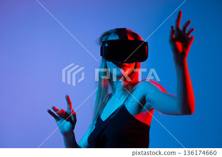 Woman in VR headset reaching upward in virtual space, neon red blue lighting, immersive technology and gesture interaction 136174660