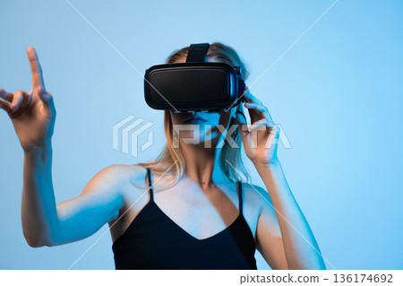 Young woman adjusting virtual reality headset while interacting with digital interface, immersive VR technology and metaverse concept 136174692