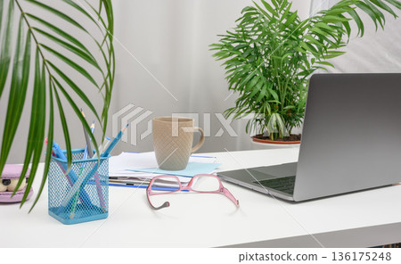 Modern workplace with open laptop, cup of coffee, pink glasses and houseplants on white table Modern workplace with open laptop, cup of coffee, pink glasses and houseplants on white table 136175248