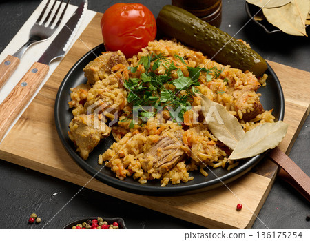Pilaf with pieces of meat and herbs served on a black plate with pickled cucumber, marinated tomato on a black background Pilaf with pieces of meat and herbs served on a black plate with pickled cucumber, marinated tomato on a black background 136175254