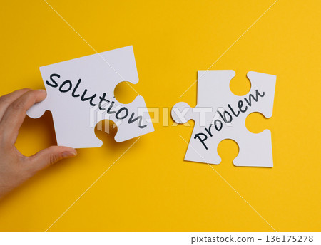 A person's hand connects two white puzzle pieces labeled solution and problem on a yellow background. A person's hand connects two white puzzle pieces labeled solution and problem on a yellow background. 136175278
