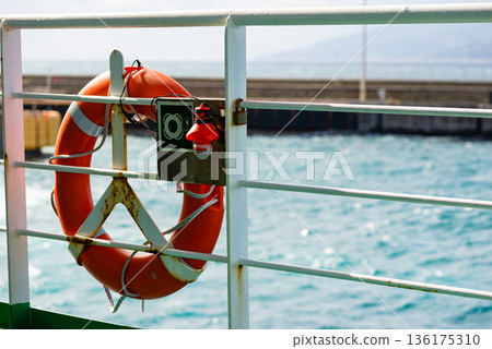 Detailed close up of orange lifebuoy on ship railing with blue sea background for maritime safety theme. 136175310