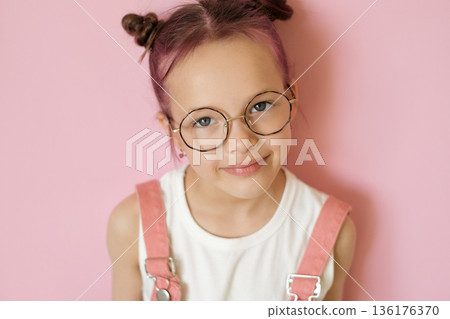 Studio portrait of a smiling girl wearing round eyeglasses on a pink background. Concept of childhood, confidence, modern eyewear and everyday vision support. 136176370