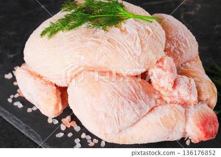 Close-up of whole frozen chicken carcass with dill and salt. Raw poultry meat covered in ice crystals on black slate board. Uncooked food ingredient ready for defrosting. High quality photo 136176852