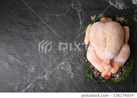 Top view of whole frozen chicken on black slate board with salt and dill. Raw poultry carcass with ice crystals on dark marble background with copy space for text. High quality photo 136176854