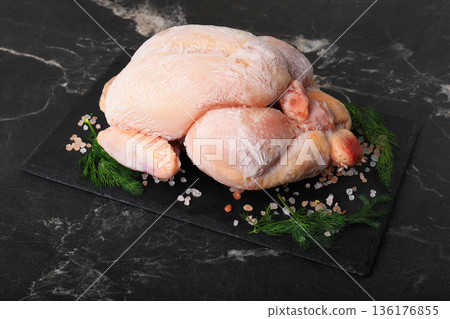 Whole frozen chicken on black slate board with salt and dill. Raw poultry carcass covered in ice crystals on dark marble background. Uncooked meat ingredient ready for defrosting. High quality photo 136176855