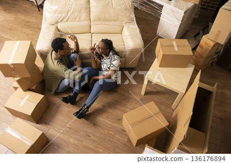 Young black couple sits on floor of new apartment surrounded by moving boxes, talking and planning decor. Spouses share conversation in unfurnished living room while unpacking belongings together. 136176894