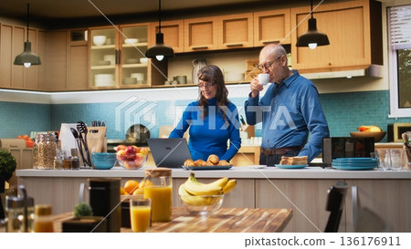 Aged husband and wife browsing laptop apps at counter in home kitchen, enjoying coffee and peaceful relaxation. Joyful lifestyle, satisfied wellbeing and relaxed moments in retirement. Camera A. 136176911