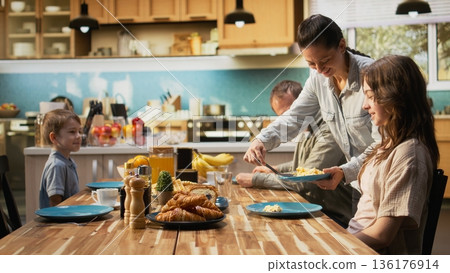 Smiling parents and two kids gathered at the breakfast table with waffles and fresh food. Kitchen scene shows morning connection, family bonding to share homemade meal together. 136176914
