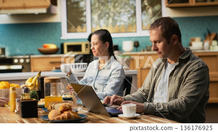 Busy father ignoring his daughter at breakfast table and focusing on work, typing information on laptop and looking preoccupied. Distant working dad with a career and pre-teen girl. 136176925