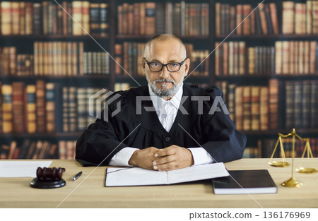 Senior judge at desk in law library with gavel, scales, and open documents 136176969