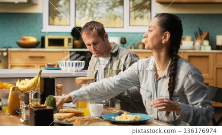 Preoccupied working dad studying work reports and documents at breakfast, ignoring his pre-teen girl and wife during family time. Father focused on papers, neglecting his daughter. 136176974