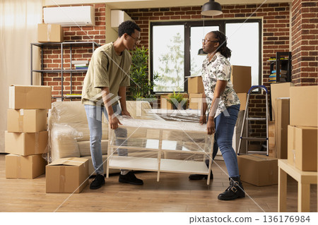 Young black couple carrying coffee table into new apartment, arranging furniture in brick wall living room. African american boyfriend and girlfriend lifting wrapped desk during moving day. Young black couple carrying coffee table into new apartment, arranging furniture in brick wall living room. African american boyfriend and girlfriend lifting wrapped desk during moving day. 136176984