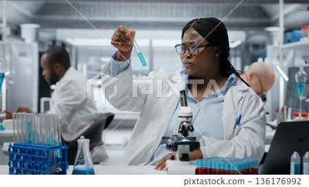 Lab materials scientist investigating blue liquid polymer properties in test tube container. Researcher testing new synthetic material durability and viscosity in lab, camera B 136176992