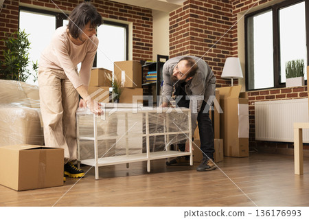 Boyfriend and girlfriend unpacking belongings in brick wall apartment, carefully placing table on floor. Caucasian newlyweds placing coffee table in living room, settling in new home. 136176993