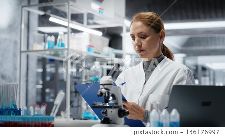 Hematology specialist in lab conducting research on blood sample, taking notes. Epidemiologist in laboratory analyzing sanguine fluid specimen in vial, writing down findings, camera B 136176997