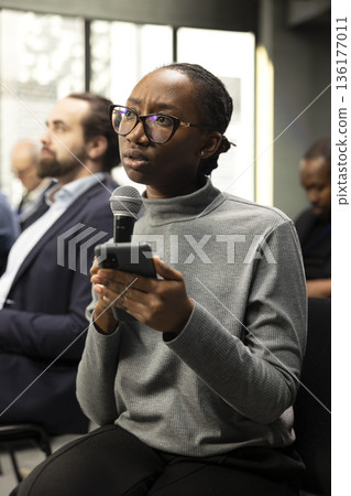 Black female journalist reading questions out loud from her phone, interacting with presenter and engaging in business discussion. Young woman writing down answers for press purposes. Black female journalist reading questions out loud from her phone, interacting with presenter and engaging in business discussion. Young woman writing down answers for press purposes. 136177011