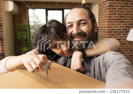 Young married couple standing with cardboard box and house keys, symbolizing fresh start and shared journey into residential property ownership. Cheerful spouses taking selfie in new apartment. Young married couple standing with cardboard box and house keys, symbolizing fresh start and shared journey into residential property ownership. Cheerful spouses taking selfie in new apartment. 136177035