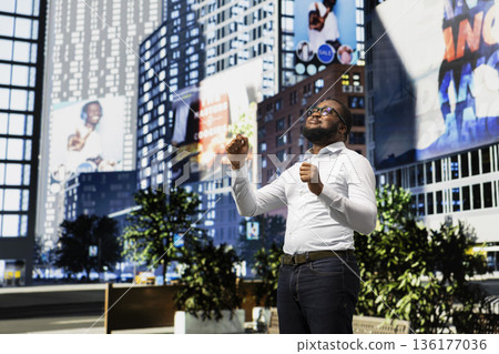 Carefree funky african american man dancing on groovy songs on the sidewalk, walking along a modern city street and enjoying music on headset. Vibrant metropolitan scenery, urbanism. Carefree funky african american man dancing on groovy songs on the sidewalk, walking along a modern city street and enjoying music on headset. Vibrant metropolitan scenery, urbanism. 136177036