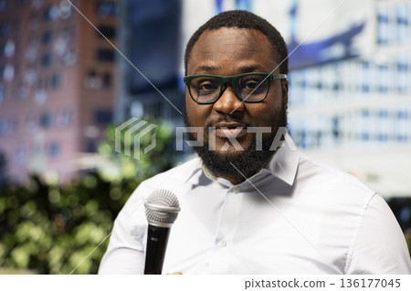 Close up of black journalist conducting on air segment in the field downtown, holding microphone and sharing breaking news information. Dedicated media career presence, city center. 136177045