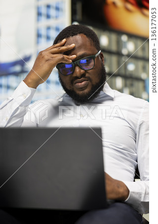 Black adult person tired and disappointed working online from park bench, being displeased and distressed with bad news on screen. Surrounded by skyscrapers in busy downtown area. 136177053
