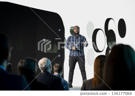 Black chief product officer introducing new smartphone with modern interface and design for marketing announcement. Mixed age audience during launch event focused on phone release. Black chief product officer introducing new smartphone with modern interface and design for marketing announcement. Mixed age audience during launch event focused on phone release. 136177068