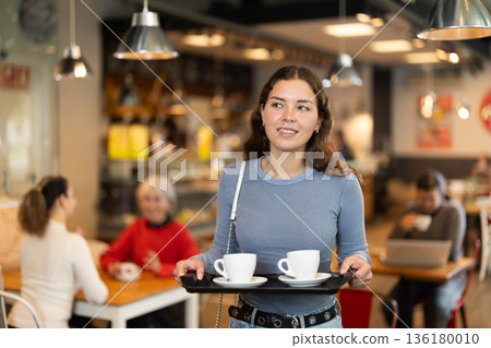 Girl visitor in self-service cafeteria carries tray with two cups of coffee. Girl visitor in self-service cafeteria carries tray with two cups of coffee. 136180010