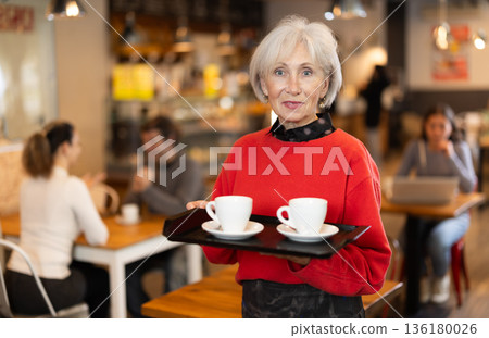 Senior woman visitor in self-service cafeteria carries tray with two cups of coffee. Senior woman visitor in self-service cafeteria carries tray with two cups of coffee. 136180026