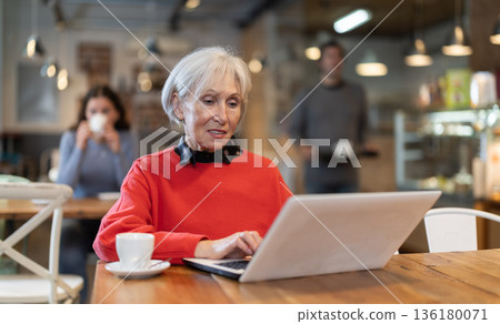 In cozy cafe hall, freelance senior woman works with laptop and drinks coffee. 136180071