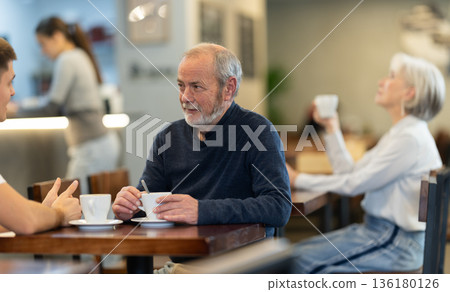 Guy in cafe sits near senior male companion and drink coffee, has nice conversation Guy in cafe sits near senior male companion and drink coffee, has nice conversation 136180126