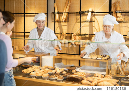 Portrait of two female bakery workers of different ages in uniform selling pastry to consumers in bakehouse Portrait of two female bakery workers of different ages in uniform selling pastry to consumers in bakehouse 136180186
