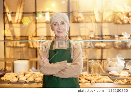 Positive mature female business owner, worker in apron standing in front of pastry stand in bakery shop. Concept of small local business Positive mature female business owner, worker in apron standing in front of pastry stand in bakery shop. Concept of small local business 136180260