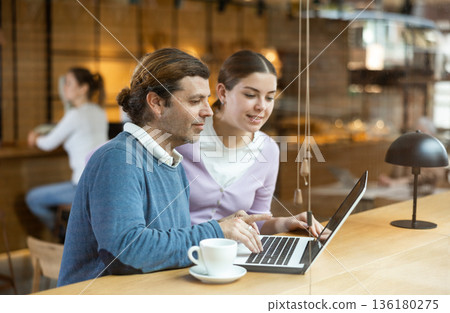 Positive European man and woman drinking and enjoying coffee while using laptop in cafe 136180275