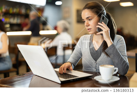 Girl freelancer with laptop spends time in cafe, talking on phone Girl freelancer with laptop spends time in cafe, talking on phone 136180313