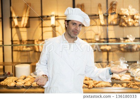 Confident adult man, bakery owner in white chef jacket and cap looking at the camera during working day in bakehouse. Cooking, culinary and bakery concept Confident adult man, bakery owner in white chef jacket and cap looking at the camera during working day in bakehouse. Cooking, culinary and bakery concept 136180333