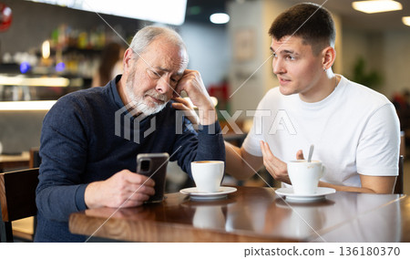 Young guy comforts mature man an upset guy while sitting at table in cafe Young guy comforts mature man an upset guy while sitting at table in cafe 136180370