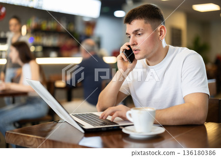 Young man with laptop talking on phone in cafe 136180386