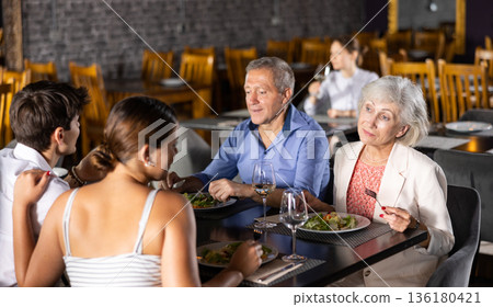 Young family and elderly couple of spouses eating in cafe celebrating event and communicating Young family and elderly couple of spouses eating in cafe celebrating event and communicating 136180421
