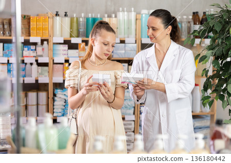 Pregnant girl looking for pills in a pharmacy. Female pharmacist helps to choose right pills 136180442