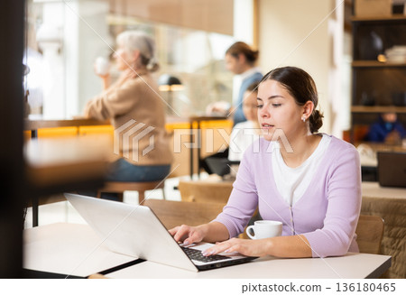 Portrait of focused young female freelancer surfing Internet on notebook while drinking tea or coffee in cafeteria Portrait of focused young female freelancer surfing Internet on notebook while drinking tea or coffee in cafeteria 136180465