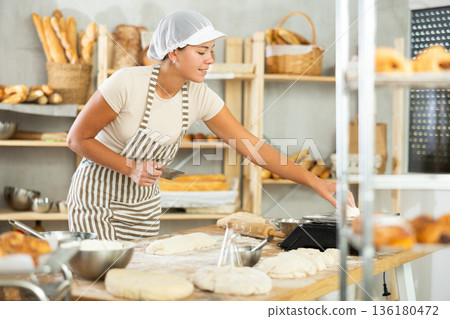 Girl baker cut small pieces of dough from large lump and put them on scales. 136180472
