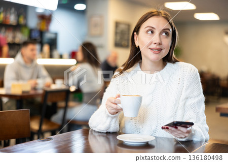 Woman with phone drinking coffee at table in cafe Woman with phone drinking coffee at table in cafe 136180498