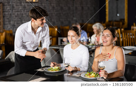 Young waiter talking to laughing female guests in restaurant Young waiter talking to laughing female guests in restaurant 136180500