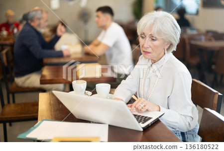 Elderly woman drinks coffee and works on a laptop in cafe interior Elderly woman drinks coffee and works on a laptop in cafe interior 136180522