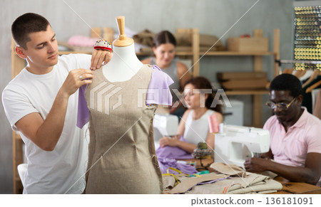 Guy student works with mannequin, others pupils sewing, working in background 136181091
