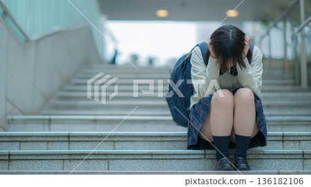 A worried Japanese female student sits on the stairs and holds her head 136182106