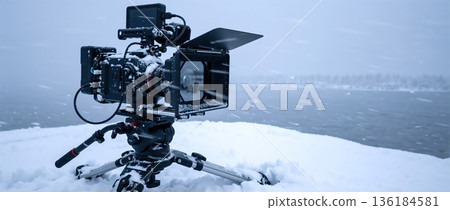 Professional cinema camera mounted on a tripod in a snowy winter landscape. Video production equipment outdoors during a blizzard with copy space 136184581