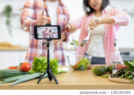 A couple is filming a cooking vlog in their kitchen, preparing a healthy salad with fresh vegetables A couple is filming a cooking vlog in their kitchen, preparing a healthy salad with fresh vegetables 136185495