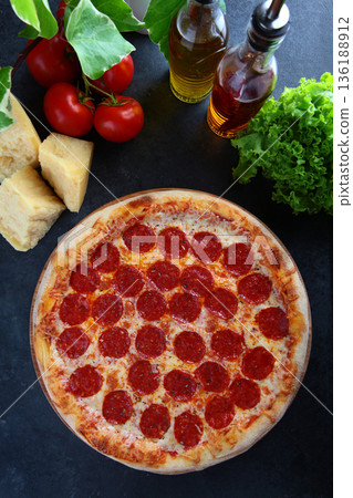 Pepperoni pizza with spicy sausage. A popular filling. Slices of hard cheese and cherry tomatoes are laid out on the table. Top view. 136188912