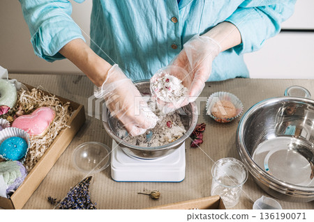 Hands mix white powder and dried botanicals in metal bowl on digital scale surrounded by finished products. Handmade bath bombs process, cosmetic chemistry at home, artisan soap making. 136190071